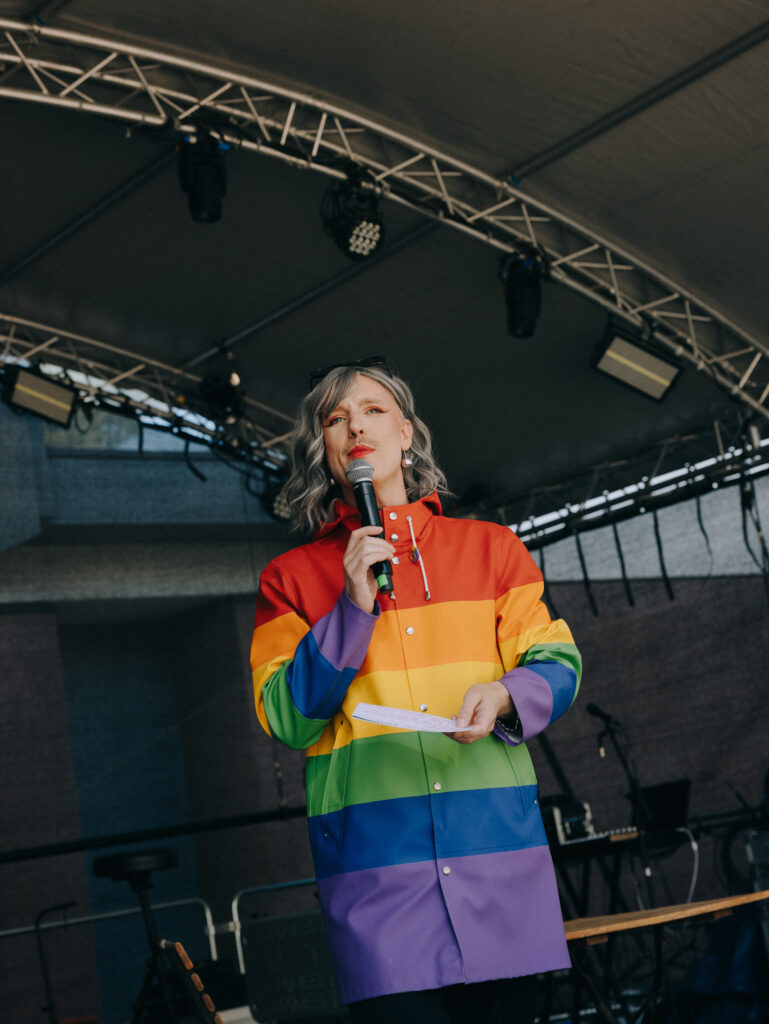 Moderator*in Janboris Ann-Kathrin Rätz in einem Regenmantel in Regenbogenfarben auf der Bühne.