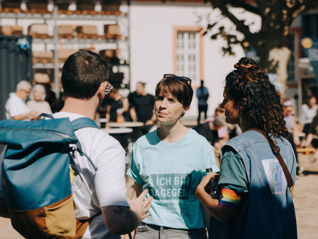 Gäste unterhalten sich beim Nie wieder ist jetzt-Minifestival im August 2026. Auf dem Shirt einer Frau steht: Ich bin dagegen, dass Menschen in Armut leben müssen.